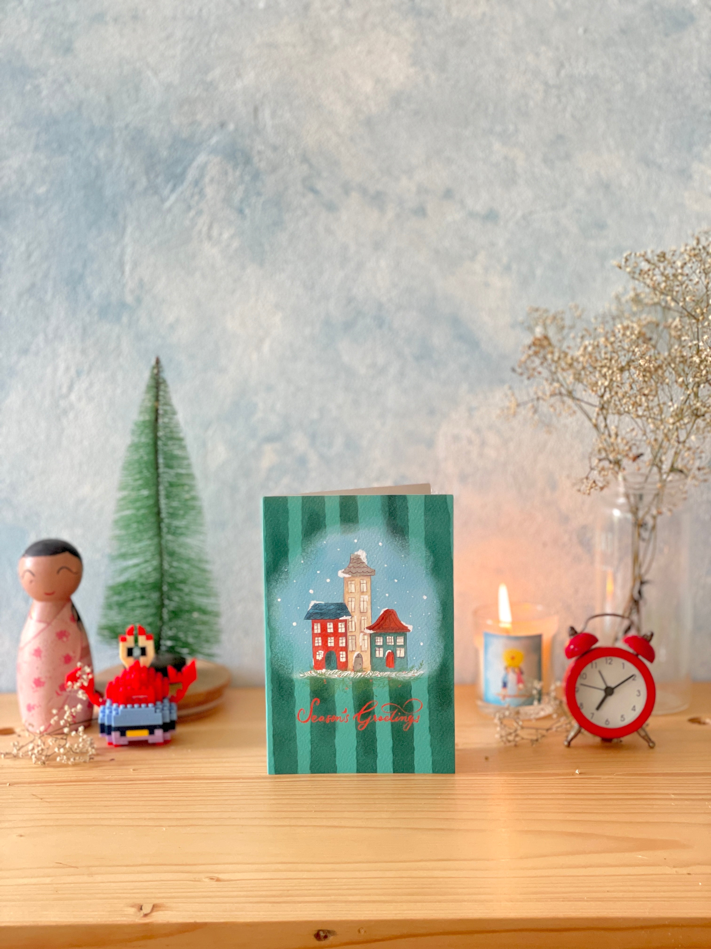Decorative card with a village scene on a wooden surface with candles and small figurines.